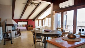 A spacious living area with a wooden dining table, comfortable seating, a ceiling fan, and large windows offering a view.