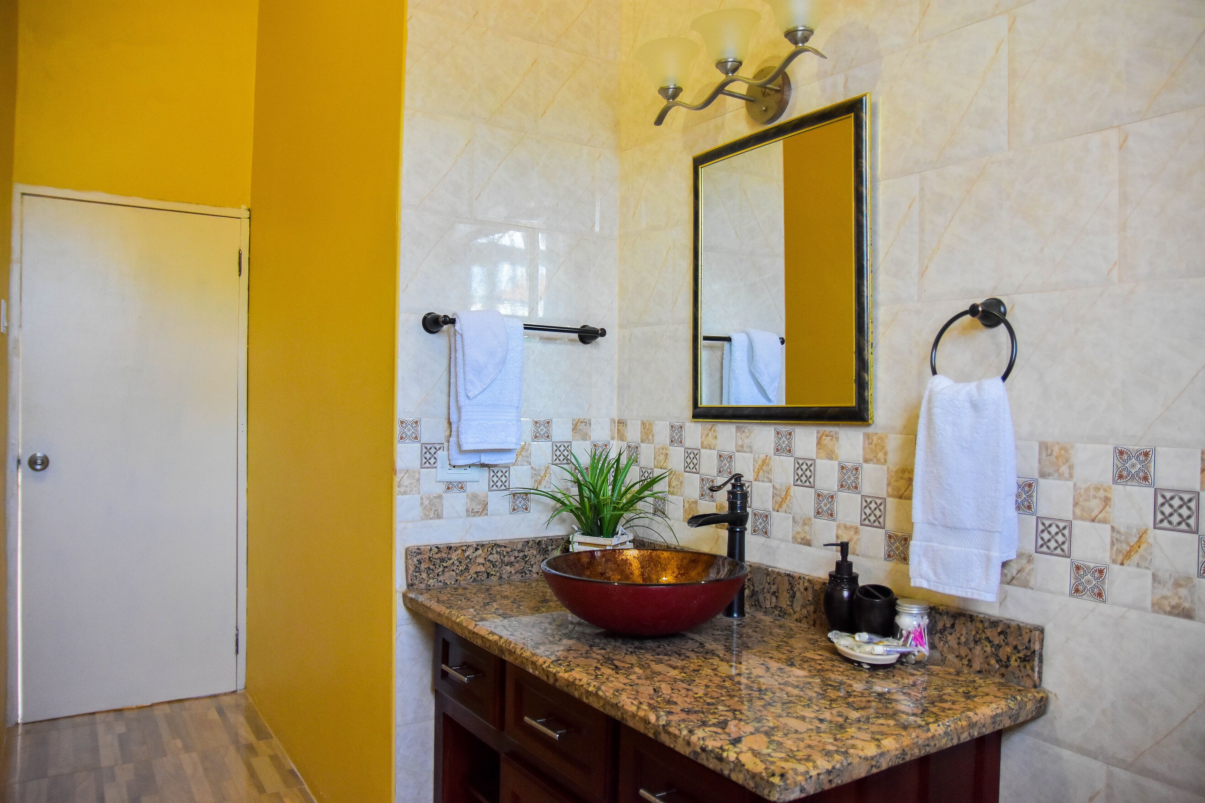 Un baño con encimera de granito, un lavamanos tipo recipiente rojo, una planta en maceta, un espejo y un perchero para toallas.