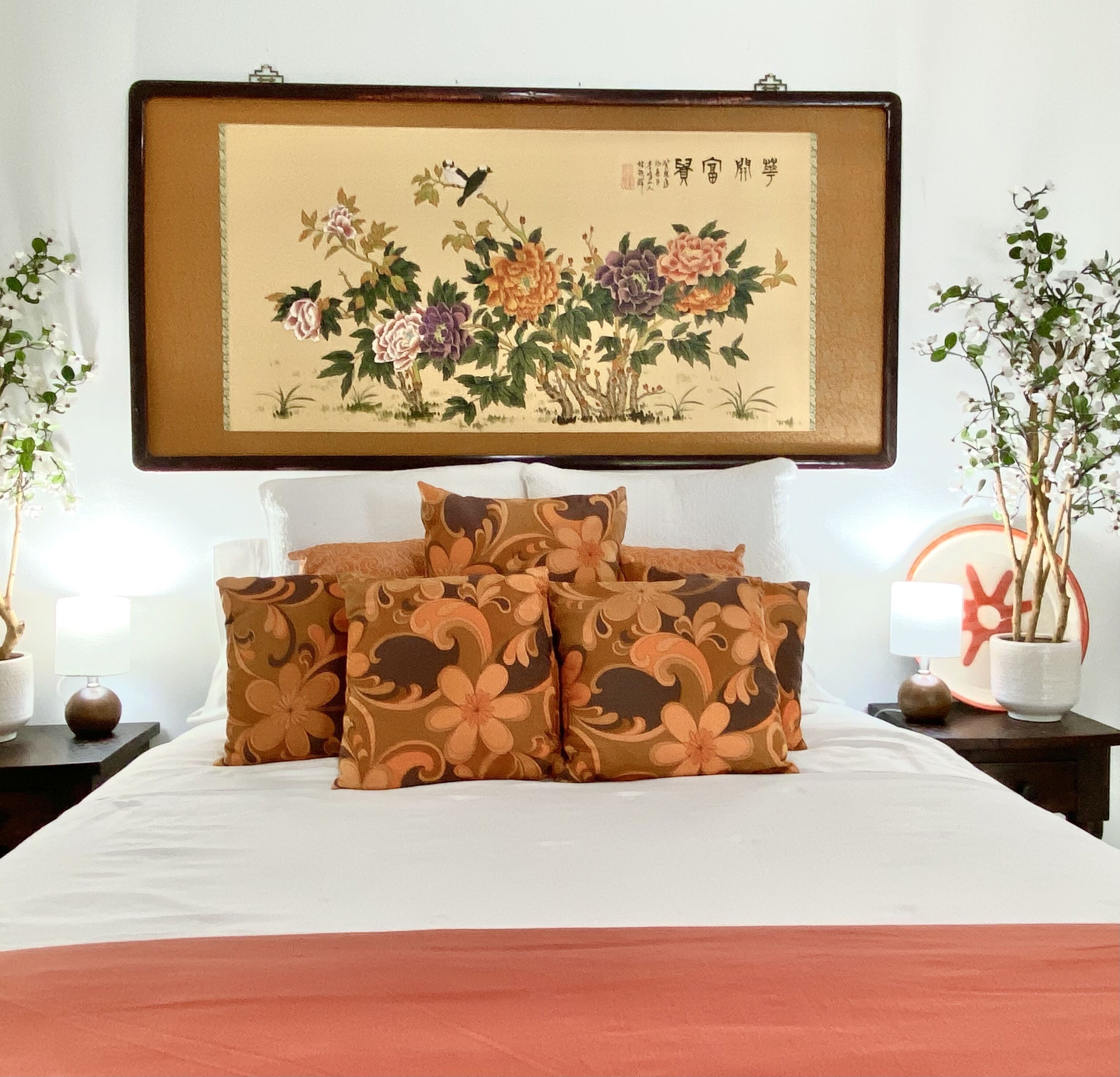 A neatly made bed with floral-patterned pillows, a framed artwork above it, and two potted plants on either side.