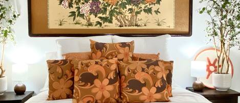 A neatly made bed with floral-patterned pillows, a framed artwork above it, and two potted plants on either side.
