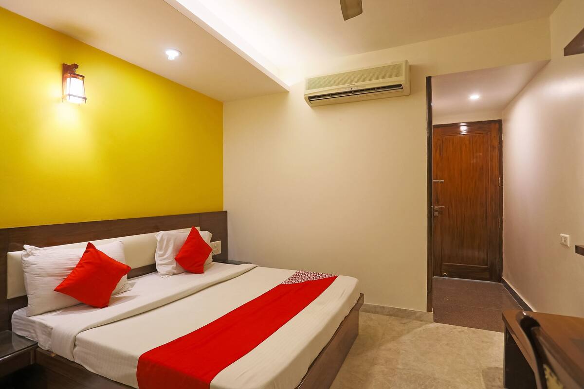 Habitación de hotel con cama, mesita de noche, colcha roja y blanca, pared amarilla y puerta de madera.
