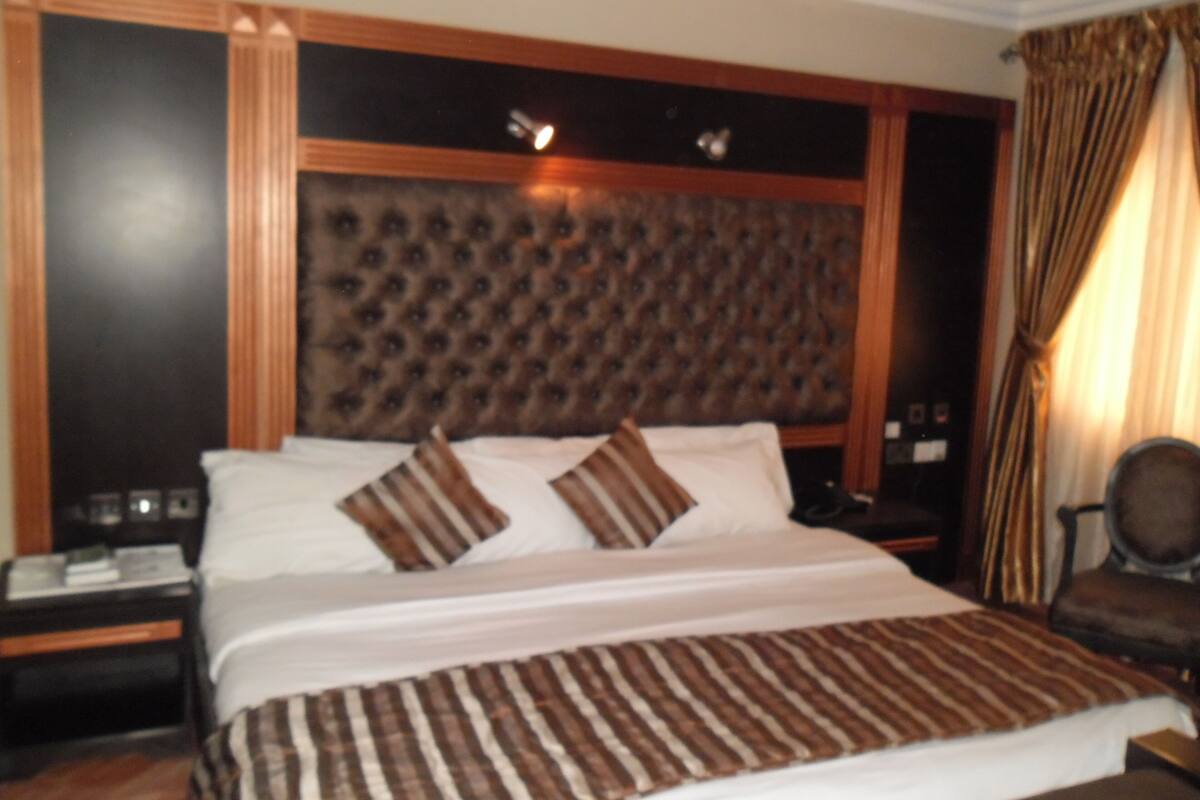 A hotel room with a large bed, a headboard with a tufted design, a nightstand with a lamp, and a chair.