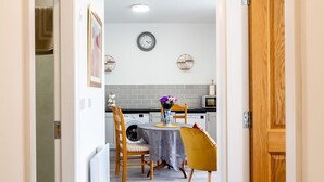 Um corredor estreito que leva a uma sala de jantar com mesa e cadeiras, um relógio na parede e uma porta de madeira à direita.