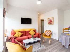 Un salon avec un canapé rouge, des fauteuils jaunes, une table basse et un téléviseur fixé au mur.