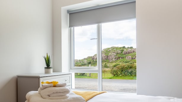 Quarto com uma janela ampla, cama com roupa de cama branca, mesa de cabeceira com uma planta e vista para uma paisagem verde.
