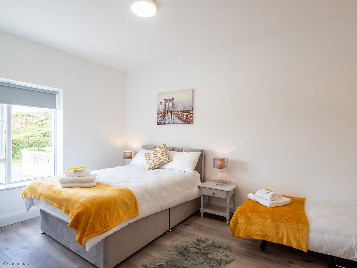 Une chambre d’hôtel moderne avec un grand lit, un plus petit lit, une table de chevet, une lampe et un tableau représentant un pont.