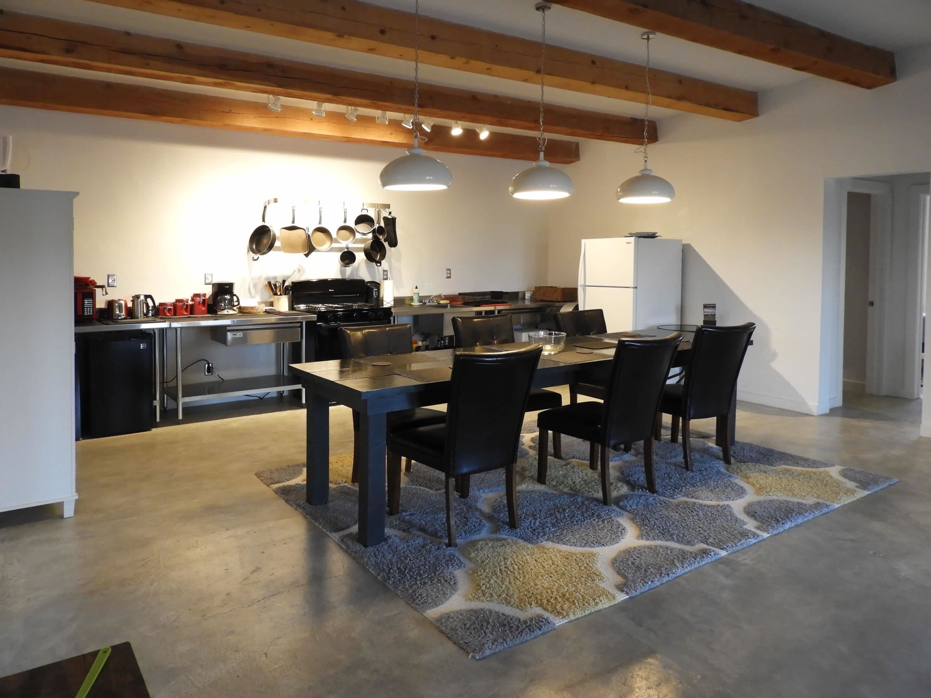 Une cuisine moderne avec un coin repas, comprenant une grande table, des chaises, des luminaires suspendus et un réfrigérateur.