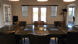 A dining area with a large table, multiple chairs, pendant lights, and a view through the door.