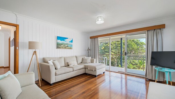 A spacious living room with a large sectional sofa, a floor lamp, a flat-screen TV, and a view of greenery through large sliding doors. - Oceanview, 6 Stewart Street (Crescent Head)