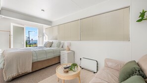 A bedroom with a bed, bedside table, lamp, radiator, and a view of the outdoors.