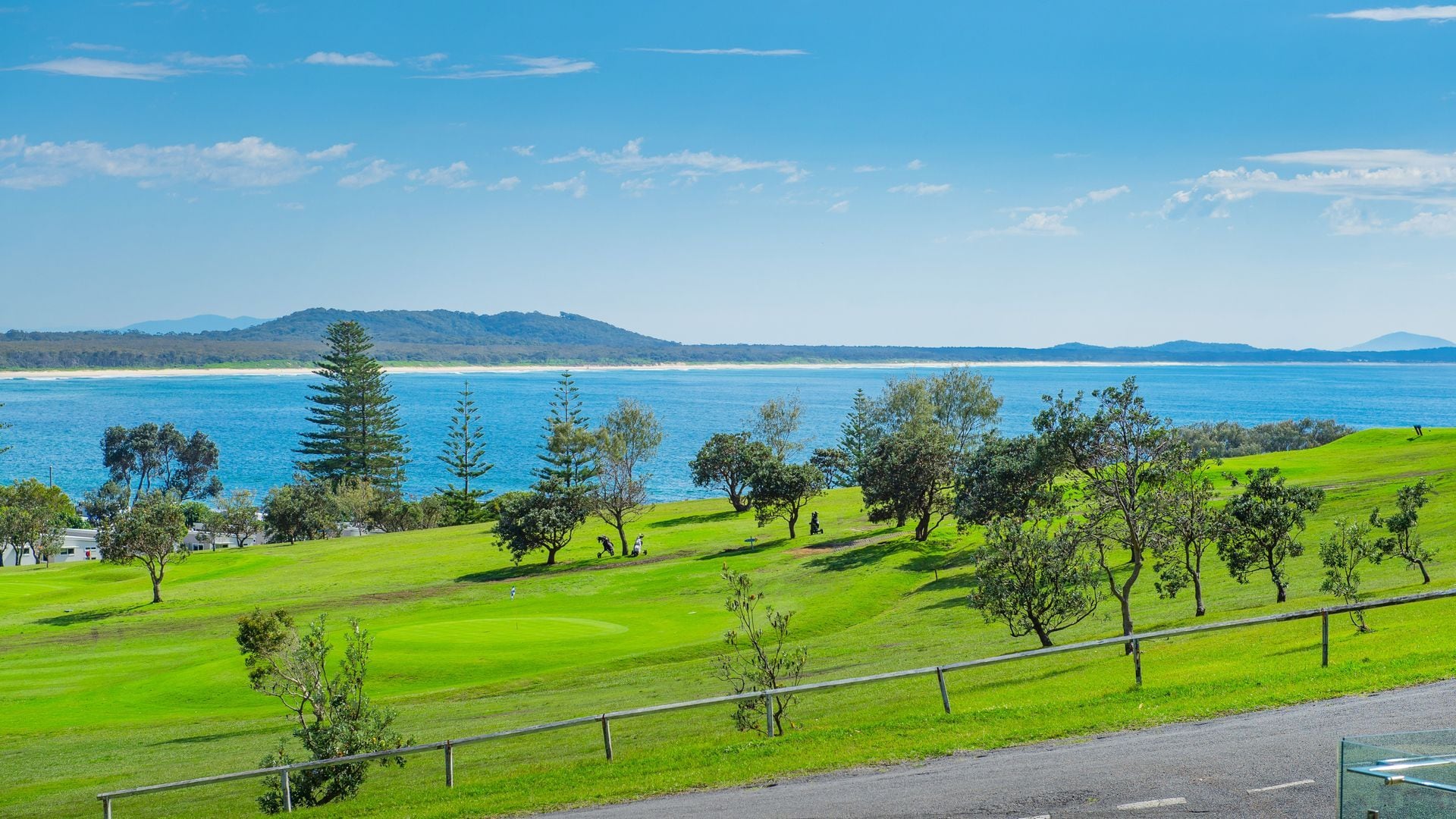 A coastal landscape with a golf course, trees, and a view of the sea and distant hills.