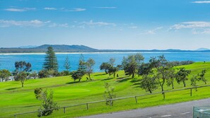 A coastal landscape with a golf course, trees, and a view of the sea and distant hills.