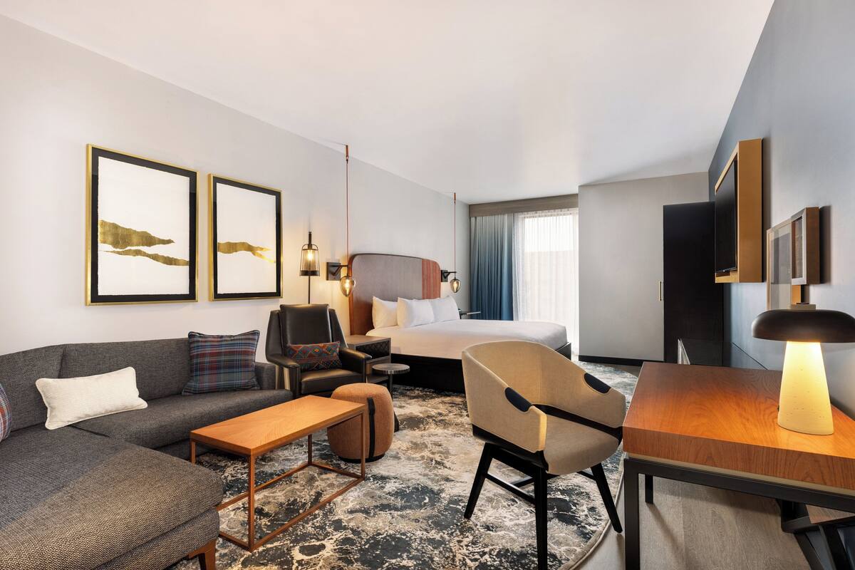 Una habitación de hotel moderna con sofá, sillón, cama y una pequeña mesa. Hay cuadros en la pared y una lámpara sobre la mesa.