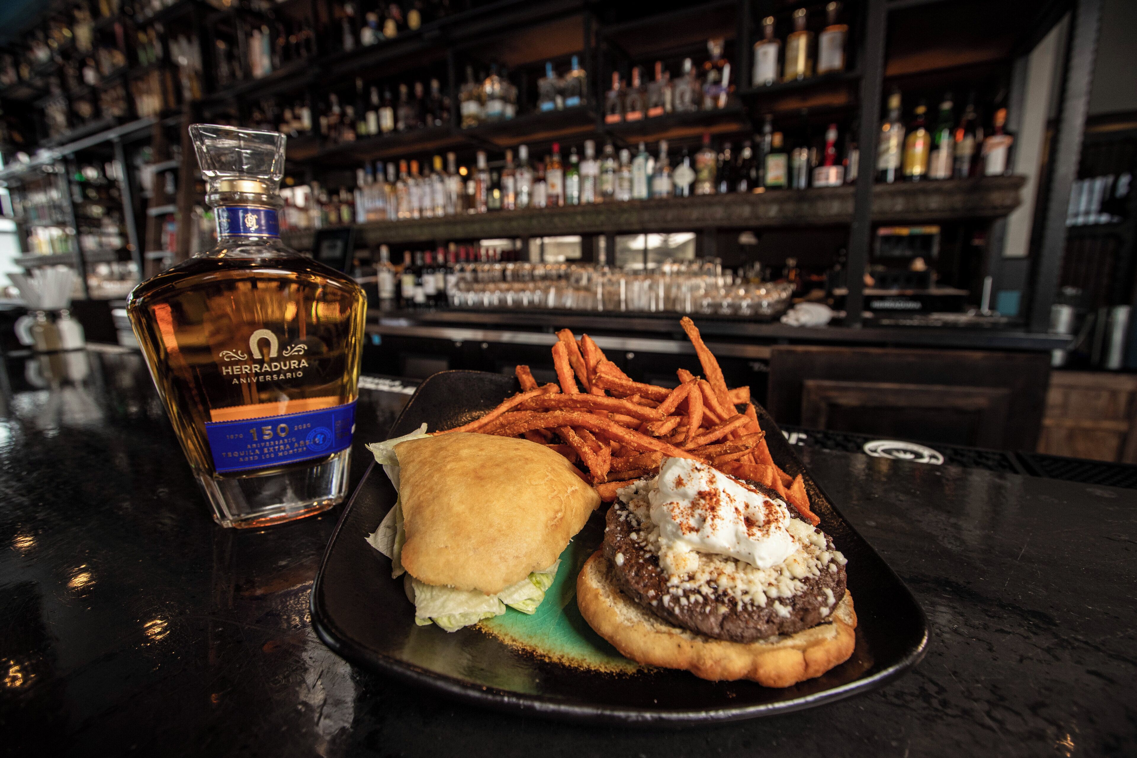 Un plato con una hamburguesa, papas fritas y una botella de tequila Herradura sobre una barra.