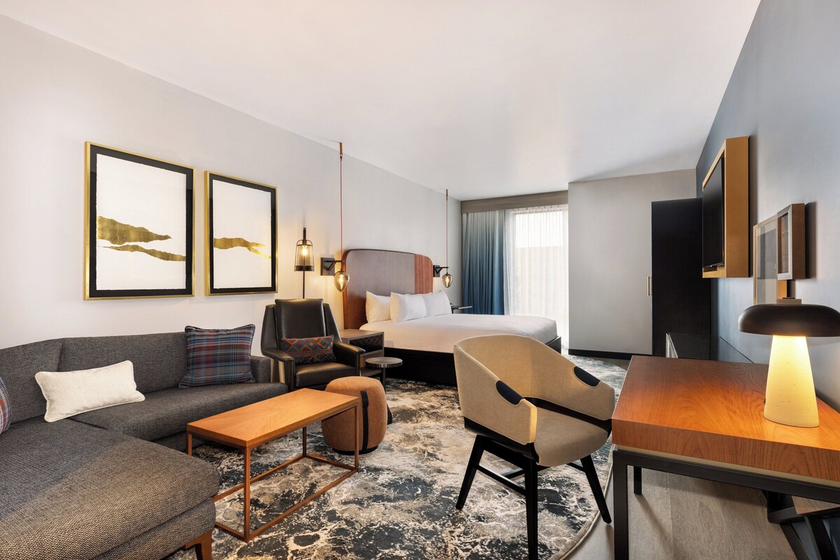 A modern hotel room with a sofa, armchair, bed, and a small table. There are framed pictures on the wall and a lamp on the table.