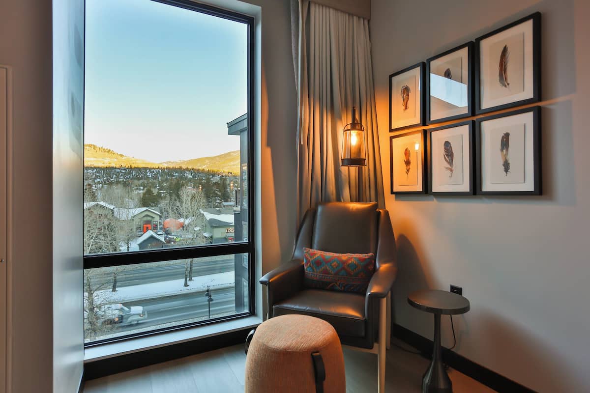 Una habitación moderna con un ventanal grande, un sillón de cuero con un cojín estampado, una pequeña mesa redonda y plumas de pájaro enmarcadas en la pared.