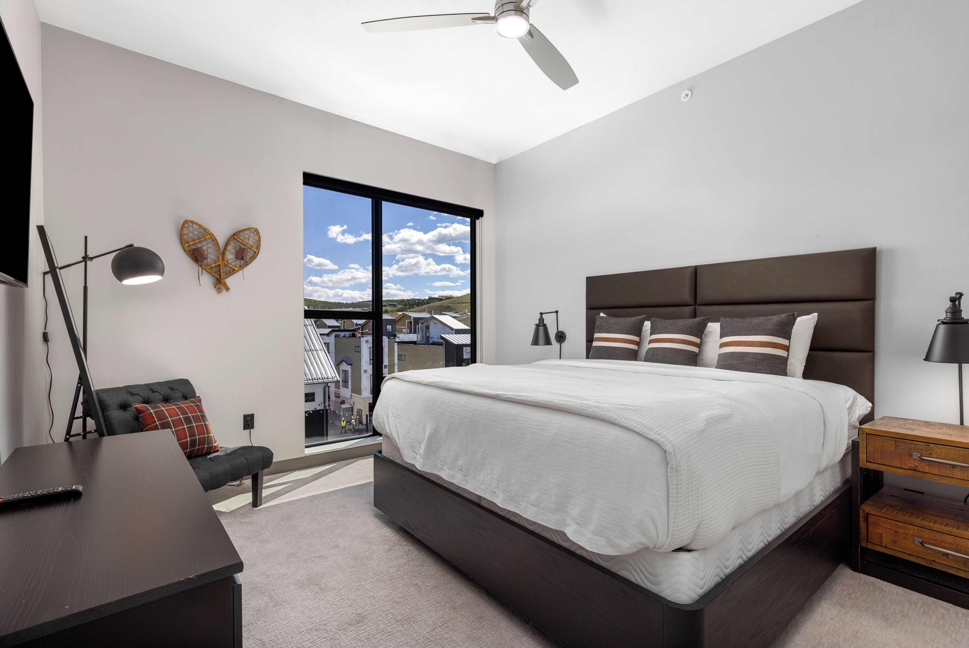 A modern bedroom with a large bed, a desk, and a view of the city through a large window.