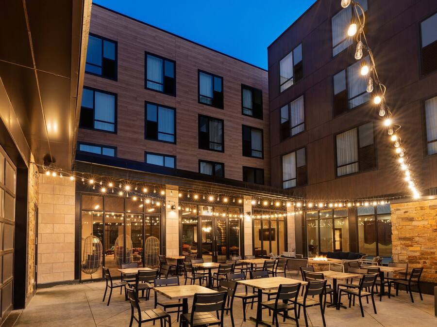 Un patio al aire libre con mesas y sillas, luces colgantes y vistas a un edificio con múltiples ventanas.