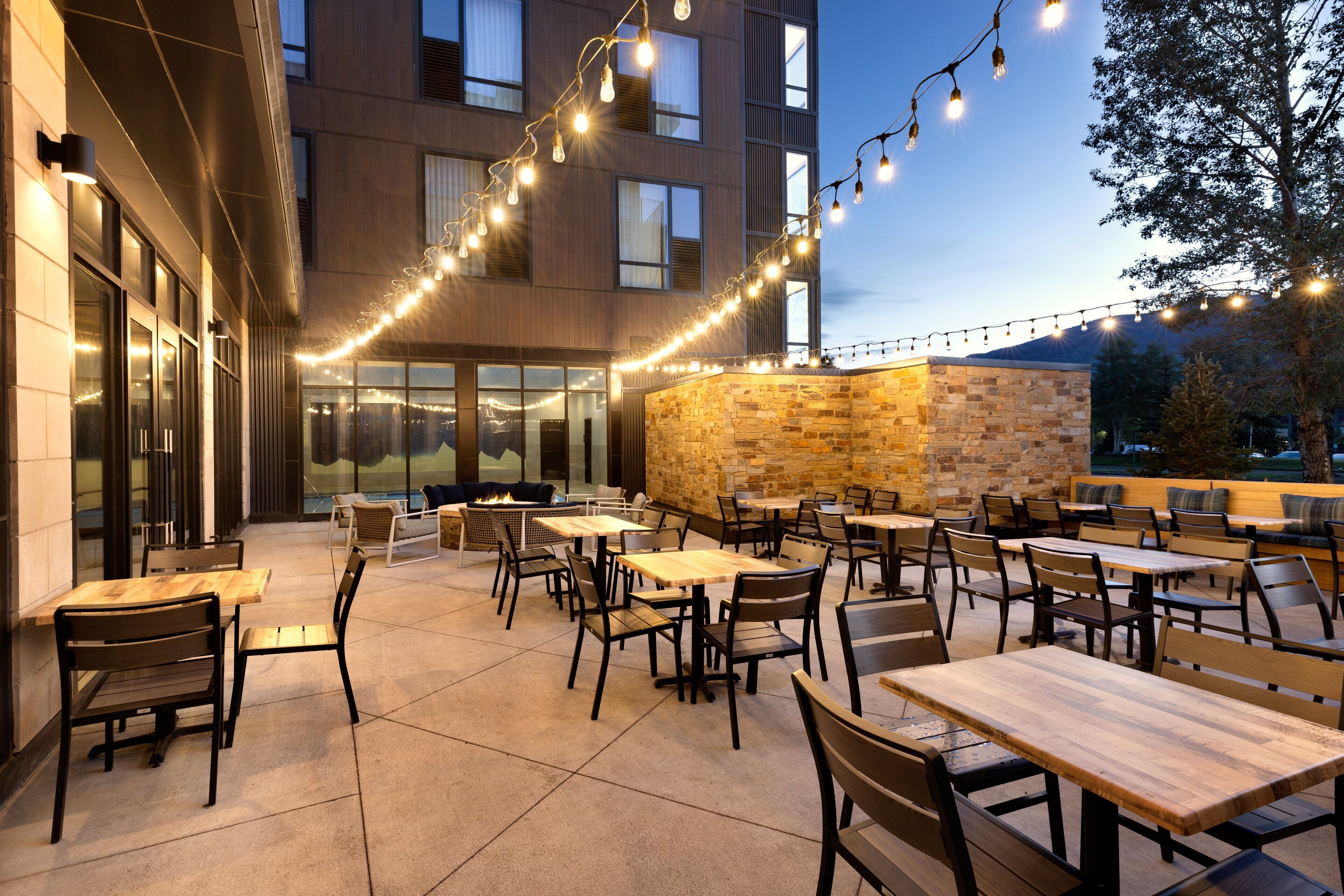 Un área de comedor al aire libre con mesas y sillas, luces colgantes y una pared de piedra.