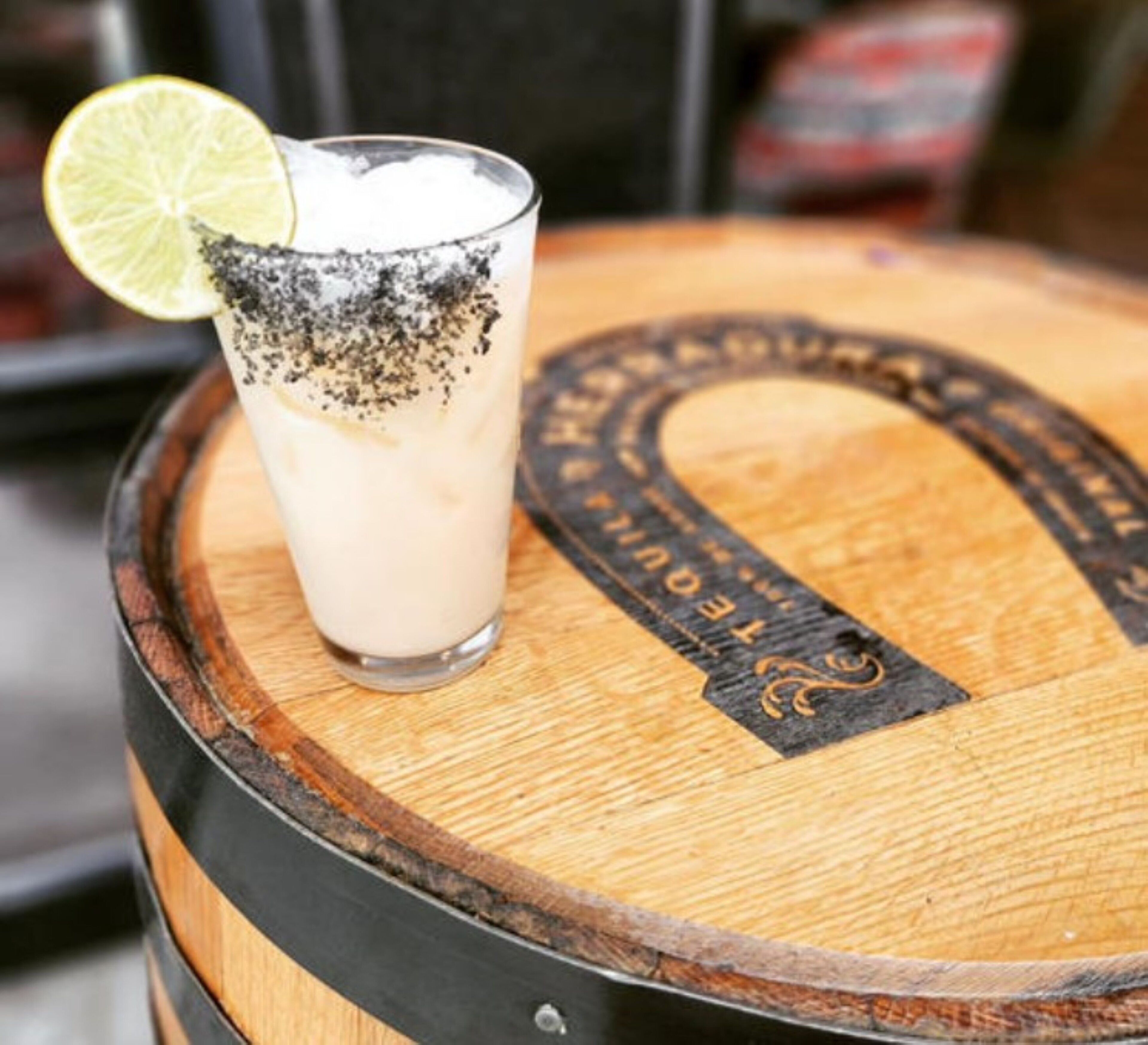 Un vaso de una bebida cremosa con una cuña de lima y un poco de polvo negro encima, sobre un barril de madera con un logotipo.