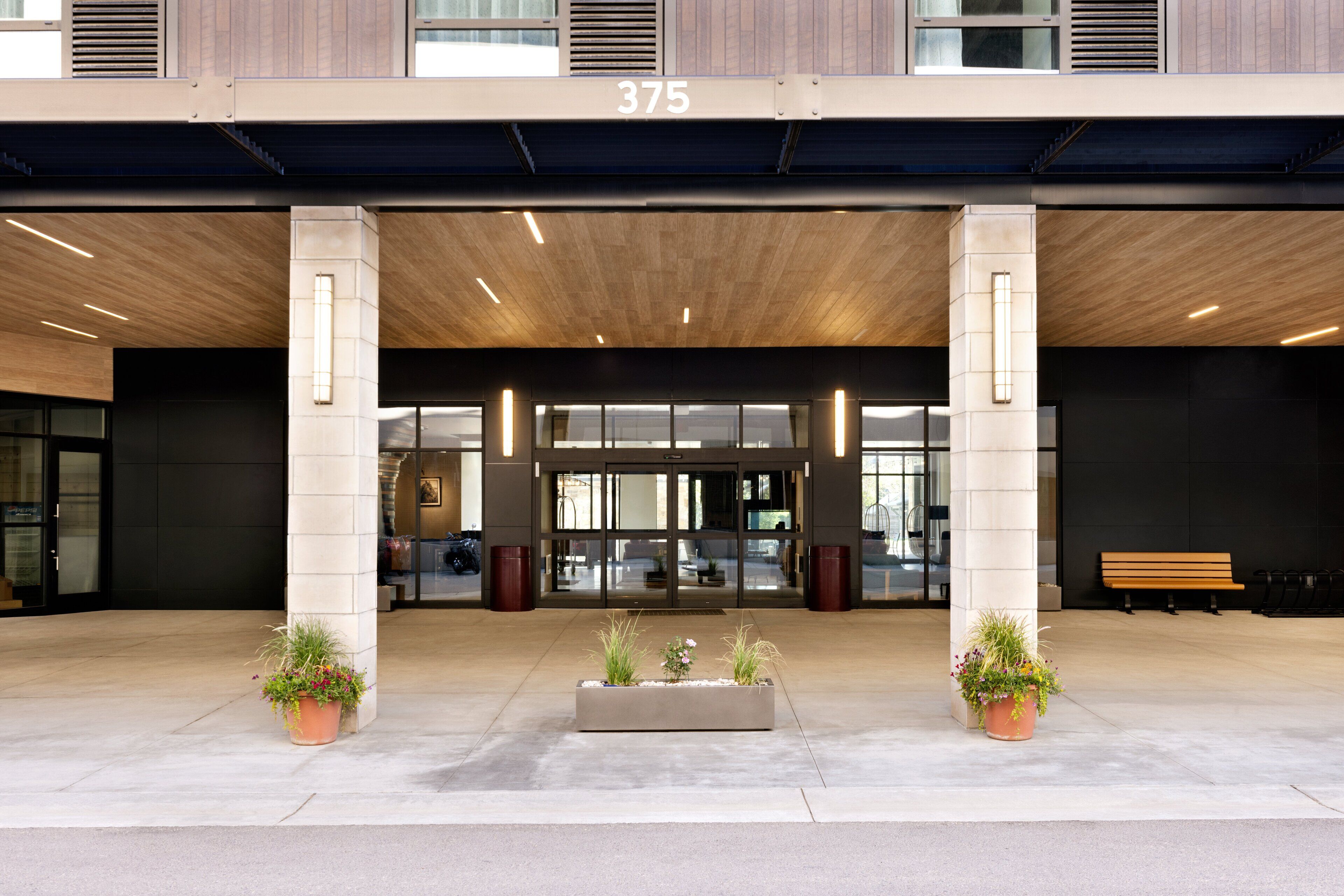 A modern building entrance with a large number "375" above the doors, two potted plants in the center, and a bench to the right.