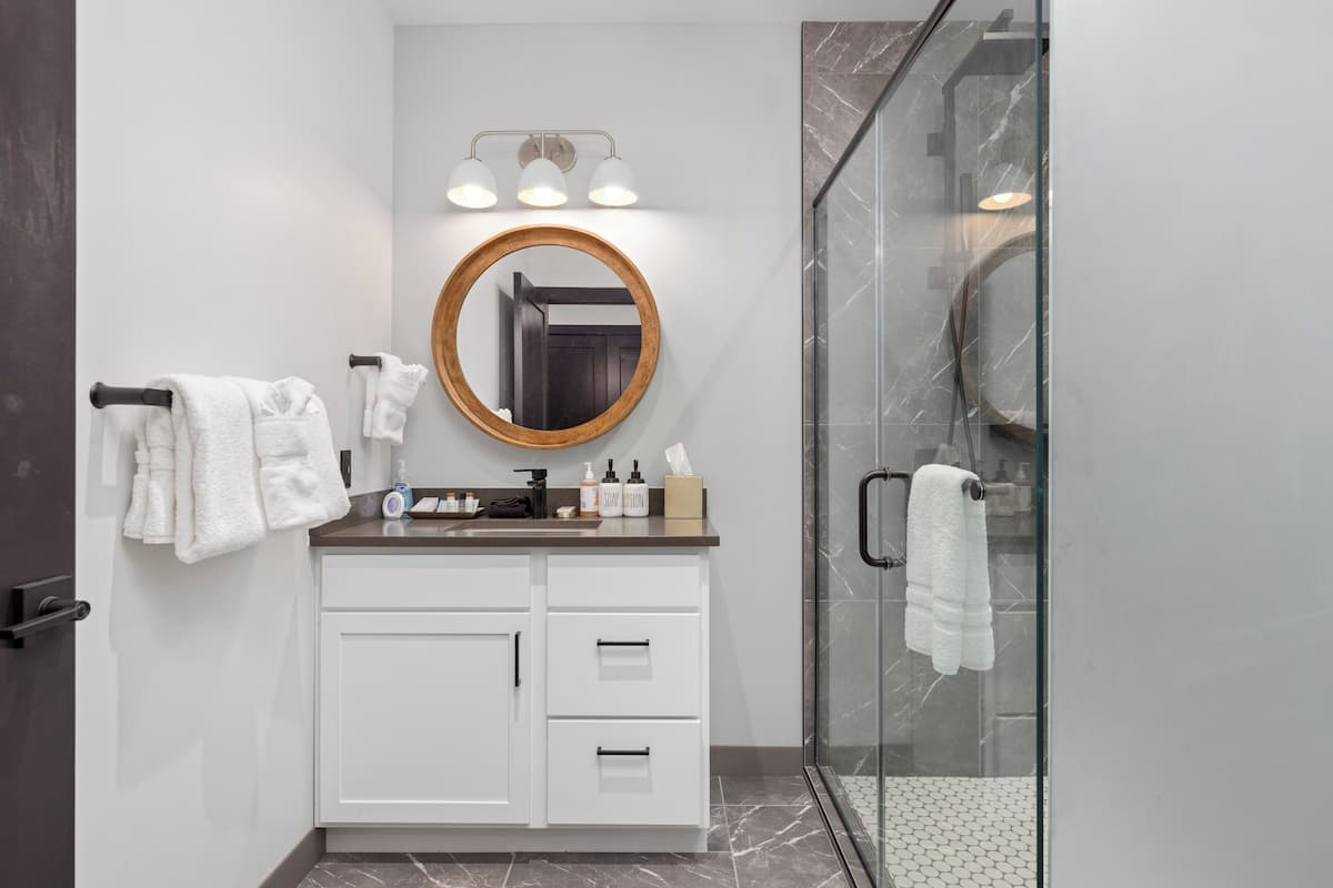 Un baño moderno con cabina de ducha de vidrio, un espejo redondo con marco de madera y un tocador blanco con encimera de mármol.