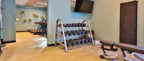 A gym with a wooden floor, a bench, dumbbells, and a TV on the wall.