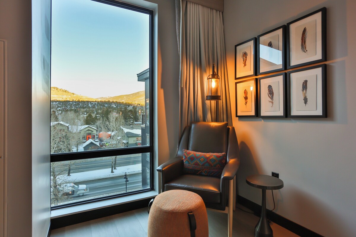 A modern room with a large window, a leather armchair with a patterned cushion, a small round table, and framed bird feathers on the wall.