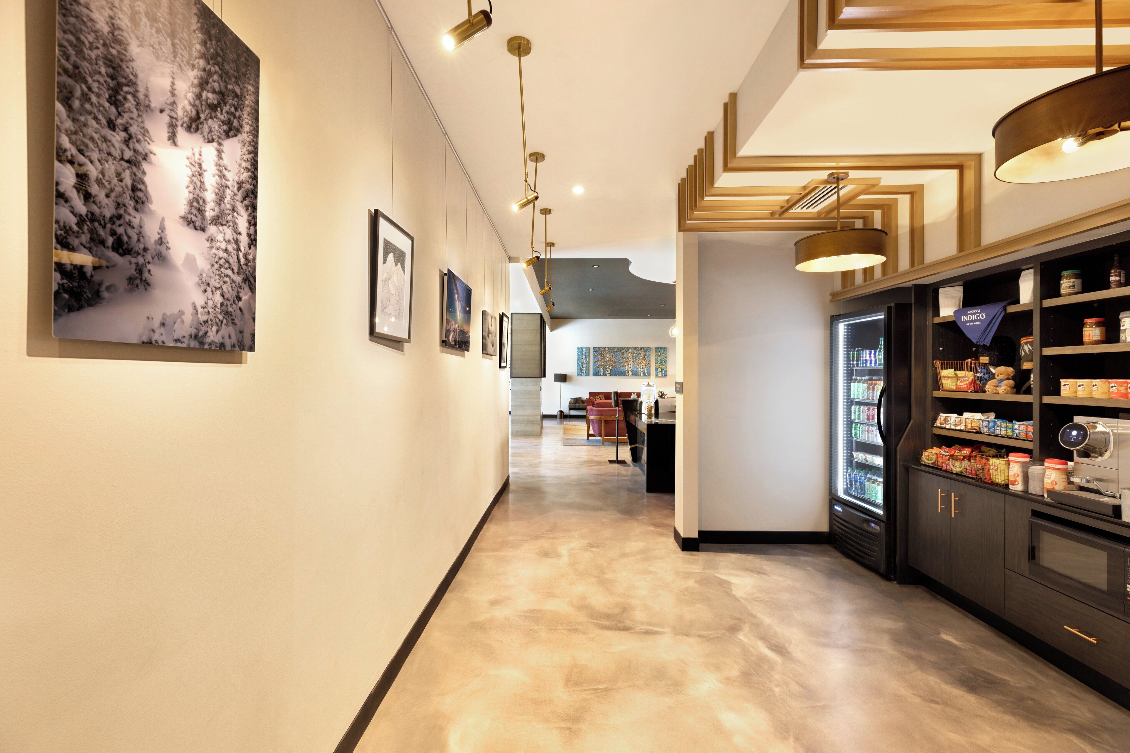 A hotel corridor with a refrigerator stocked with beverages, framed artwork on the walls, and a seating area in the distance.