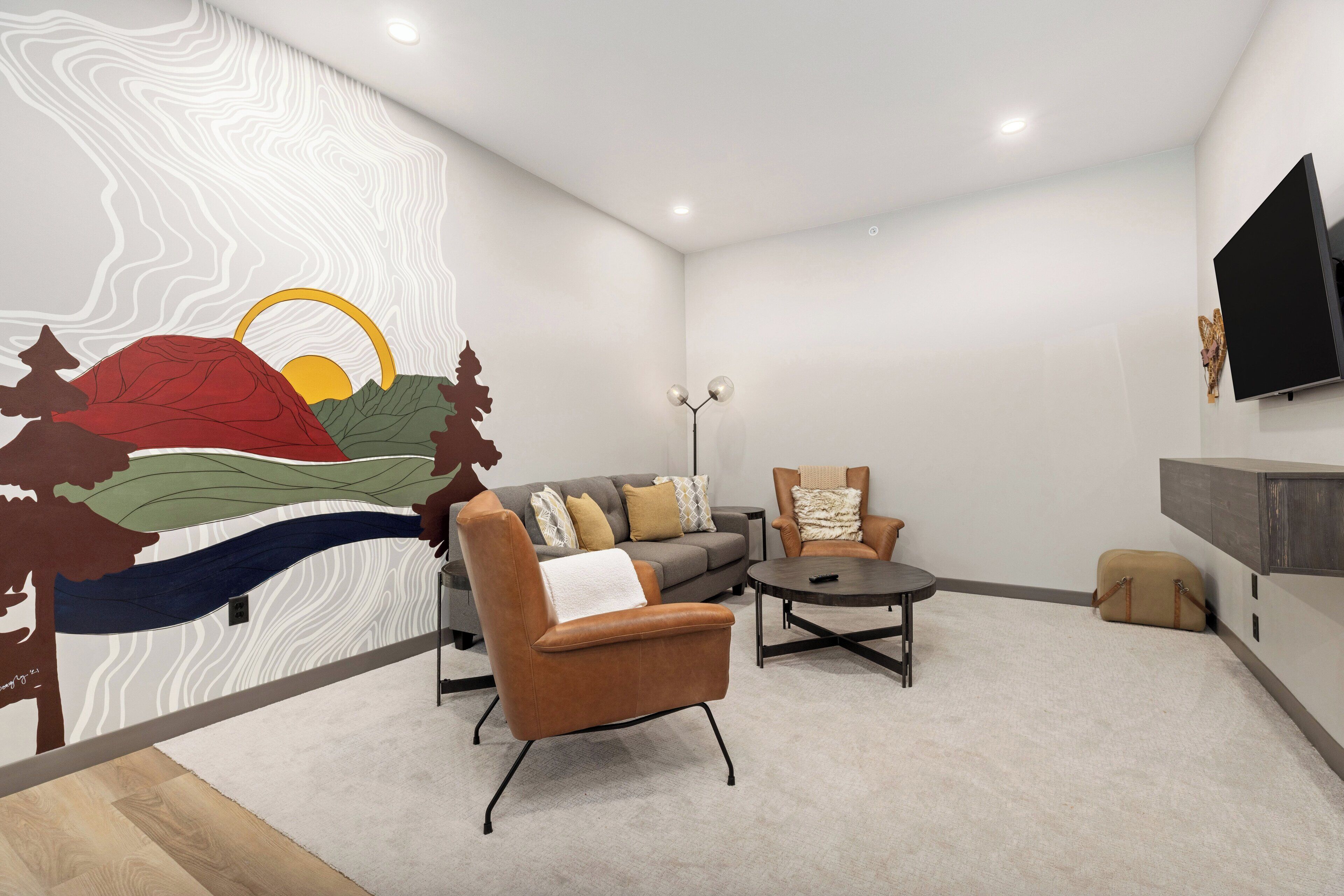 A modern living room with a sofa, armchair, and coffee table, a flat-screen TV mounted on the wall, and a decorative wall mural.