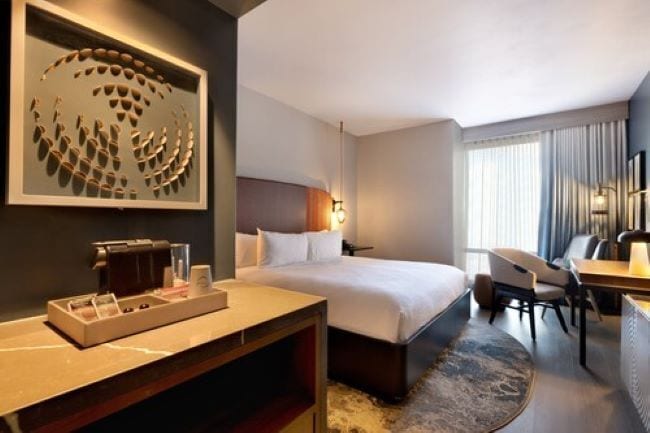 A hotel room with a bed, a desk, a chair, a coffee machine, and a decorative wall piece.