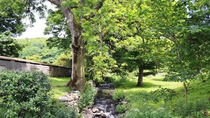 A serene outdoor scene with a small stream, lush greenery, and a stone wall. - Character former shepherds cottage for couples or young families. (near Bootle)