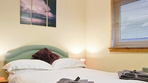 En pænt redt seng med puder, en sengegavl og en sengebordlampe. Der er håndklæder lagt sammen på sengen, og et vindue med rullegardiner.