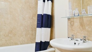 Et badeværelse med et hvidt badekar, en hvid vask med to haner, og en glashylde med toiletartikler.