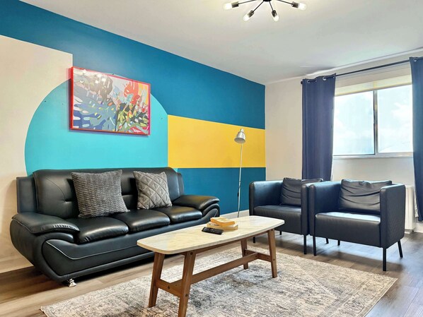 A modern living room with a black leather sofa, a wooden coffee table, and a colorful abstract painting on the wall.
