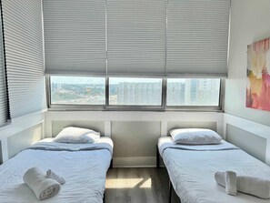 A hotel room with two beds, a window with blinds, a view of buildings, and a painting on the wall.
