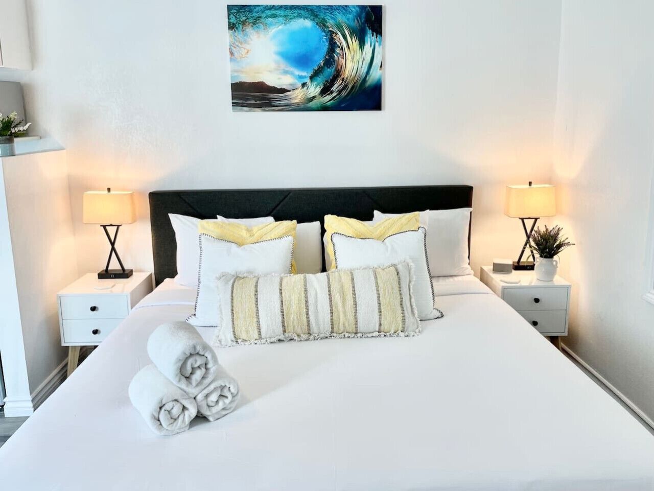A neatly made bed with white bedding, two pillows, and rolled towels. There are two bedside tables with lamps and a wall-mounted artwork above the bed.