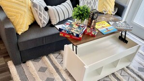 Ein modernes Wohnzimmer mit einem grauen Sofa, gelb-weißen Kissen mit Muster, einem Couchtisch mit Gesellschaftsspielen, einer kleinen Pflanze und einer weißen Aufbewahrungsanlage.