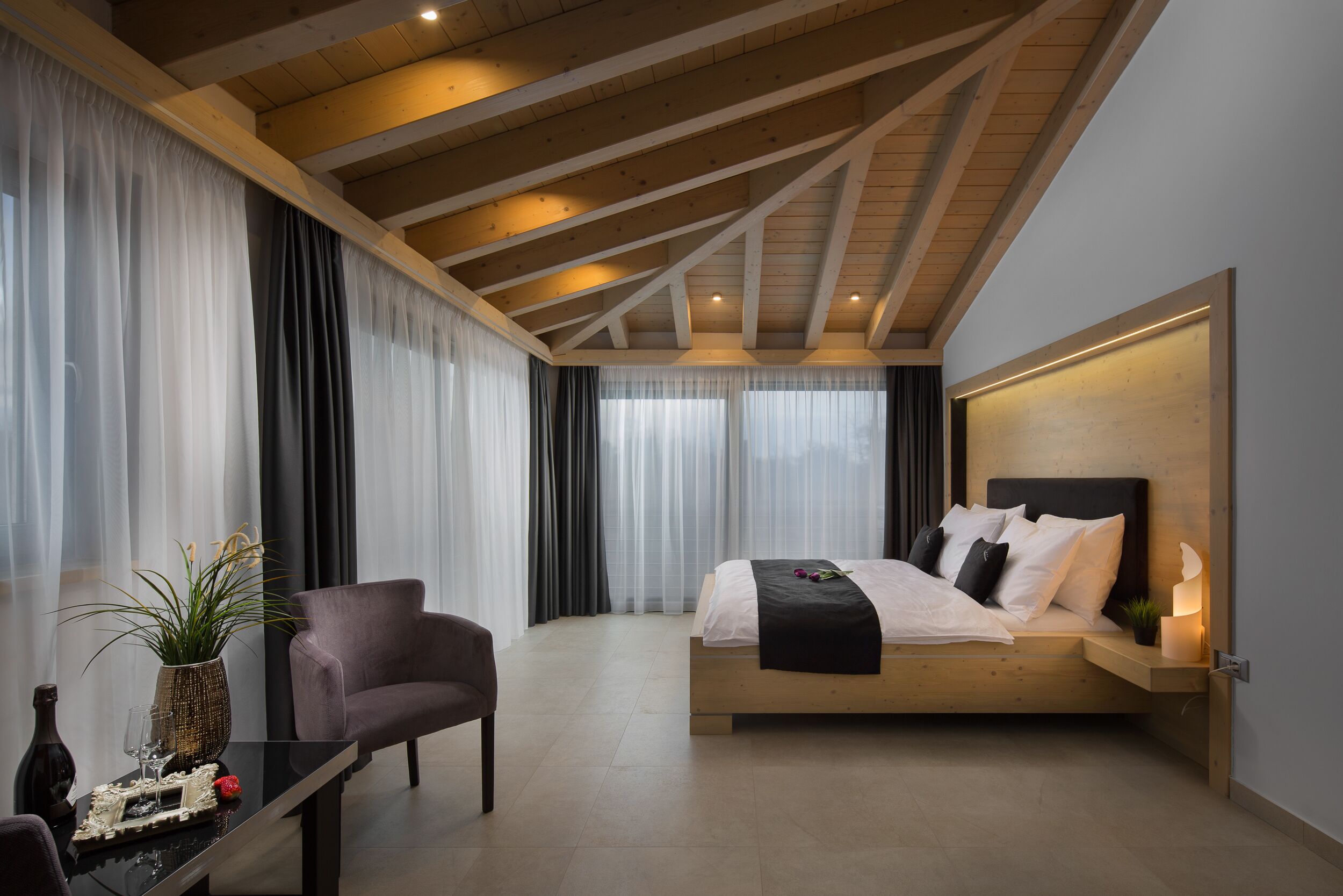 Un dormitorio moderno con techo de madera, una cama con ropa de cama blanca, una mesita de noche con un florero y una bandeja, y una silla.
