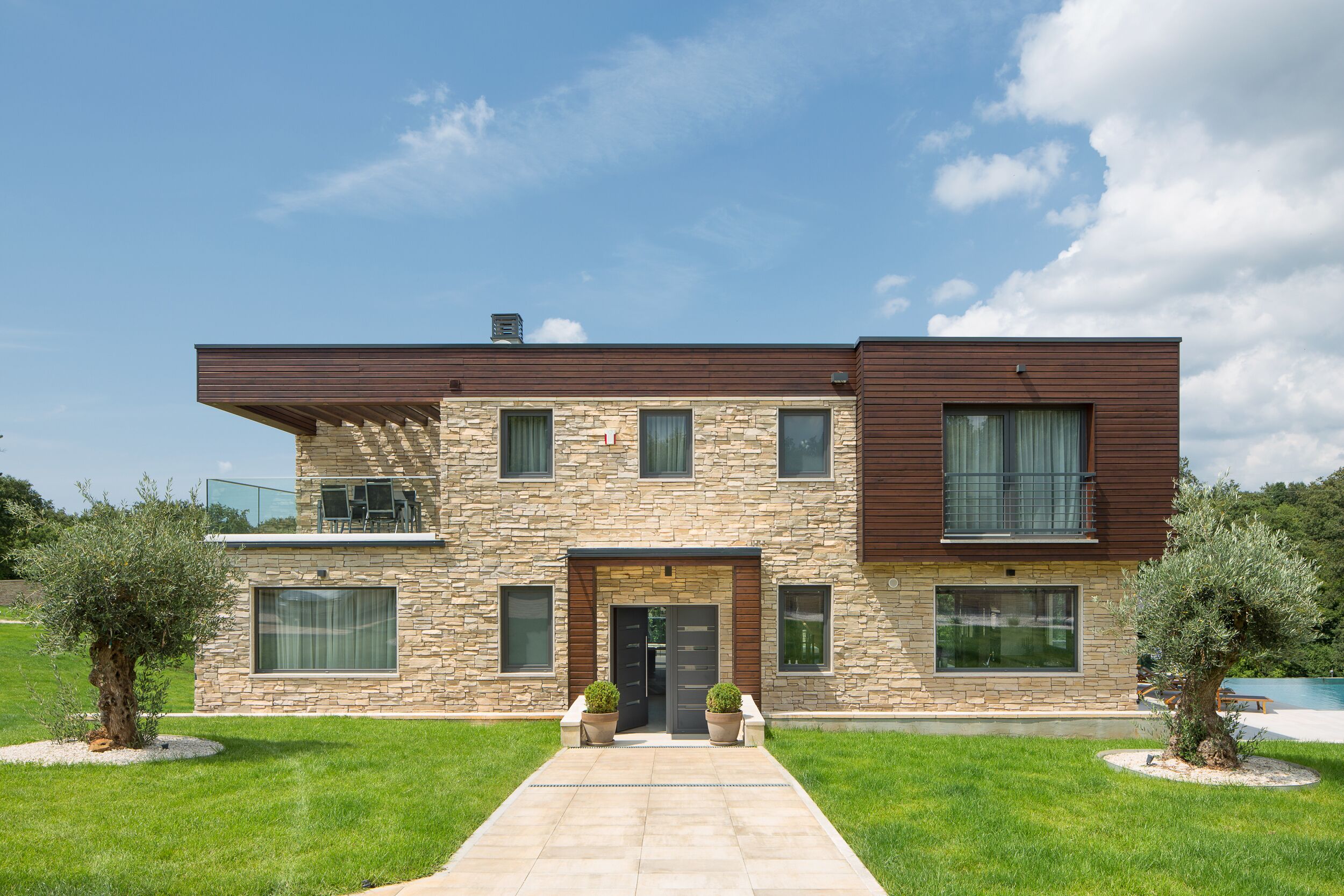 Une maison moderne à deux étages, dotée d’une façade en pierre, d’un toit plat et d’une entrée avec une porte et des fenêtres.