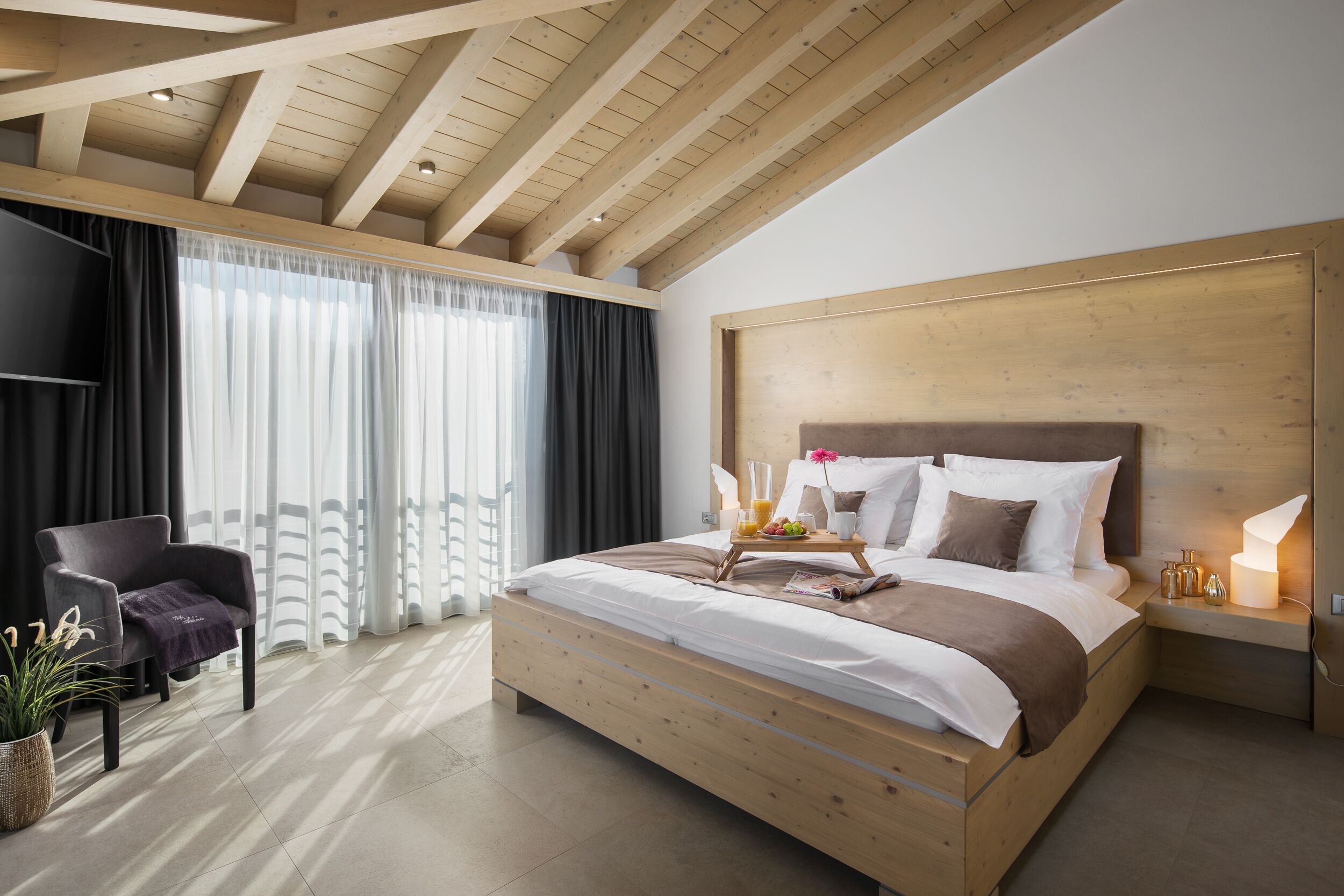 Quarto moderno com teto de madeira, uma cama grande, mesa de cabeceira com abajur e uma cadeira.