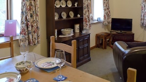 A dining area with a wooden table set for two, a television, and a bookshelf with decorative plates.