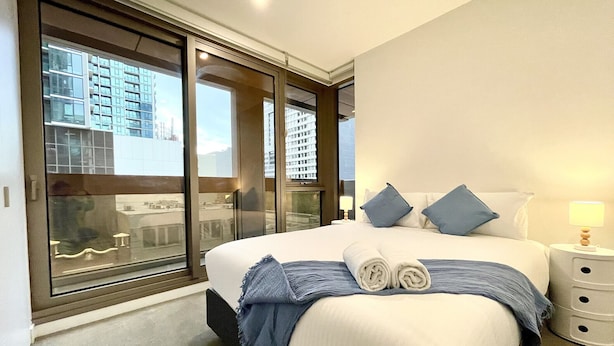 Una habitación de hotel moderna con una cama grande, vistas a través de ventanales de piso a techo y una mesita de noche con una lámpara.