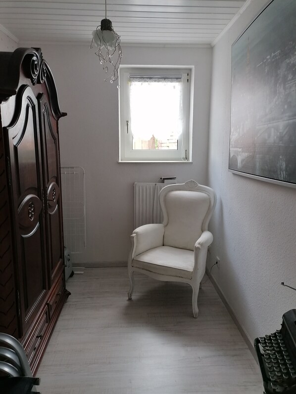 Ein Zimmer mit Holzschrank, weißem Sessel, Heizkörper, Fenster mit Blick und einem an der Wand montierten Projektor.