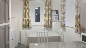 Una habitación pequeña y limpia con una mesa y sillas blancas, un radiador, una ventana con cortinas y una lámpara fijada a la pared.
