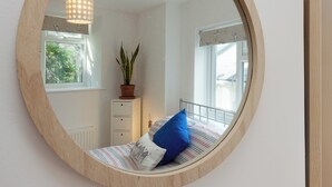 Une chambre avec un miroir rond, un lit avec une literie à rayures, une plante en pot et une commode.