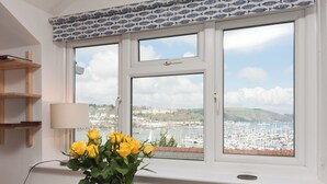 Une pièce avec vue sur un port de plaisance, un vase rempli de fleurs jaunes posé sur une table et une fenêtre ornée d’un rideau à motifs.