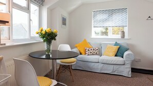 Un salon moderne avec une table ronde, deux chaises blanches, un canapé gris agrémenté de coussins jaunes et bleus, et une fenêtre donnant sur un paysage aux motifs variés.