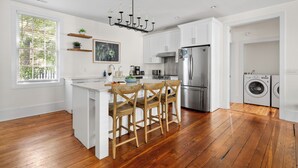 A modern kitchen with white cabinets, wooden flooring, a central island with bar stools, stainless steel appliances, and a large window with blinds.