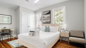 A bedroom with a large bed, a desk, a chair, a nightstand, and a window with blinds.